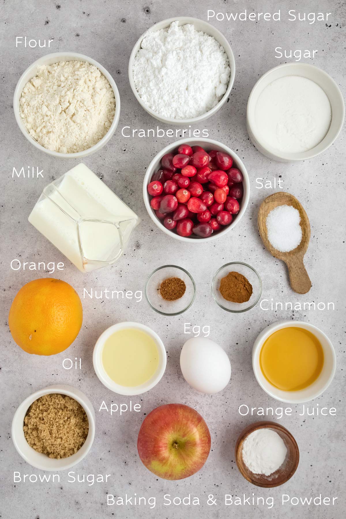 A variety of labeled baking ingredients arranged on a gray surface, perfect for making easy sheetpan pancakes, including flour, powdered sugar, cranberries, milk, orange, spices, oil, egg, apple, brown sugar, and leavening agents.