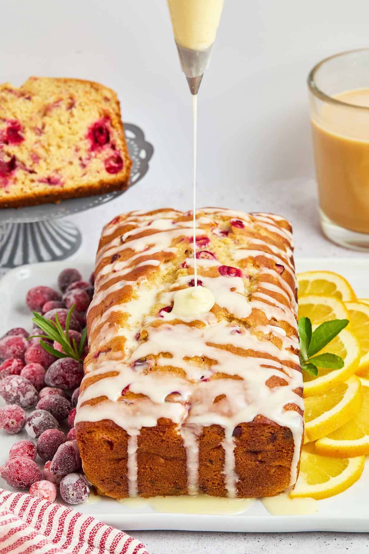 A loaf of cranberry lemon bread is being drizzled with icing, surrounded by sliced lemons and sugared cranberries, while a glass of beige drink and a plate of sheet pan pancakes sit in the background.