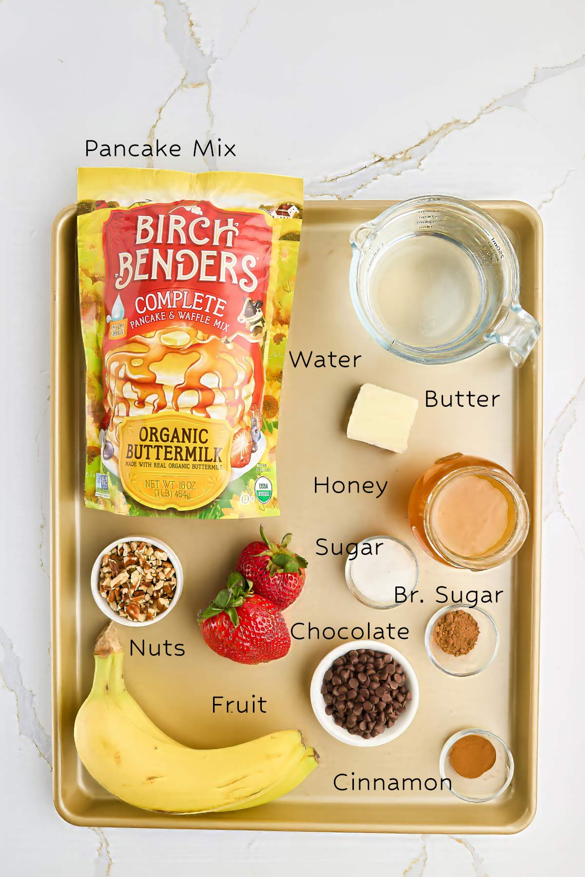 A tray with pancake mix, water, butter, honey, sugar, brown sugar, chocolate chips, cinnamon, chopped nuts, strawberries, and a banana, all labeled.
