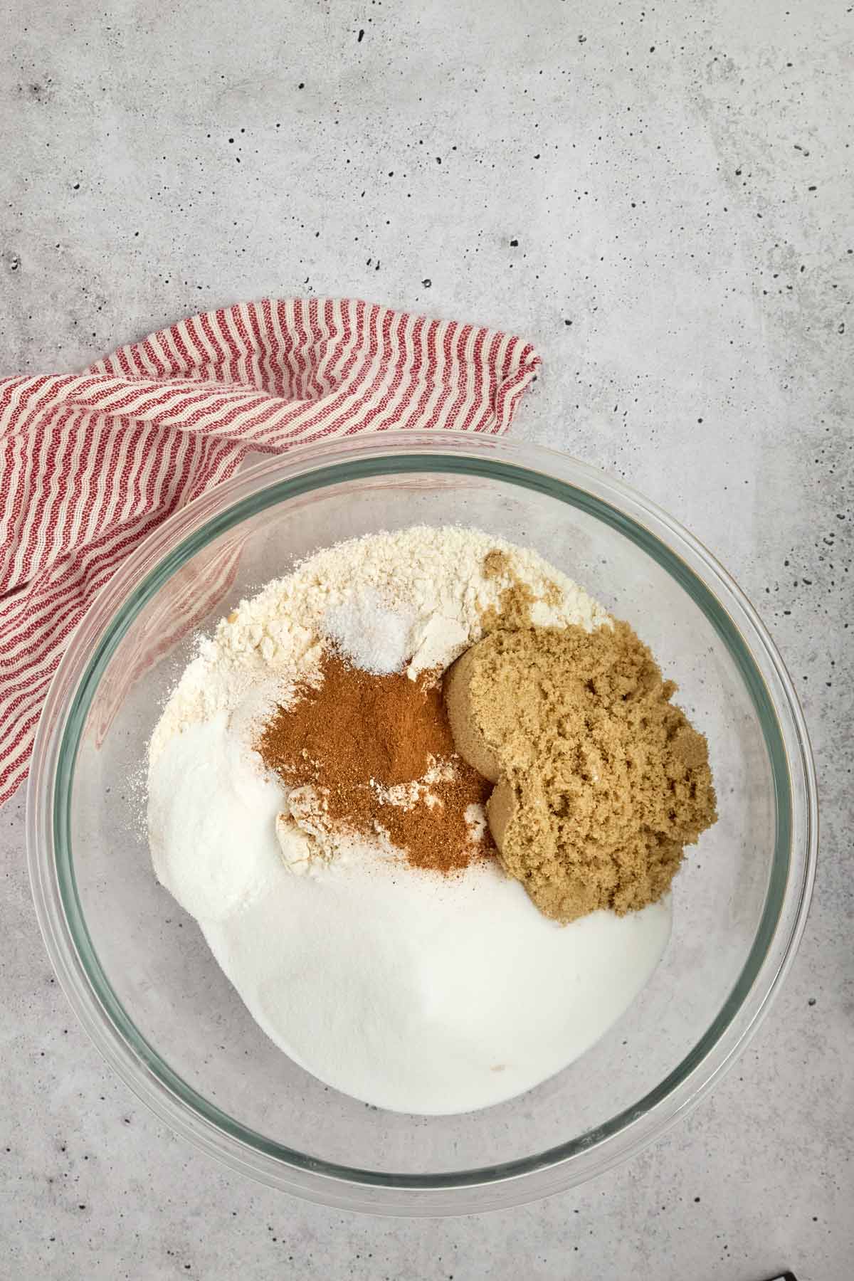 A glass bowl with flour, white sugar, brown sugar, and cinnamon sits on a light gray surface next to a red and white striped cloth-perfect ingredients for an easy sheetpan pancake recipe.