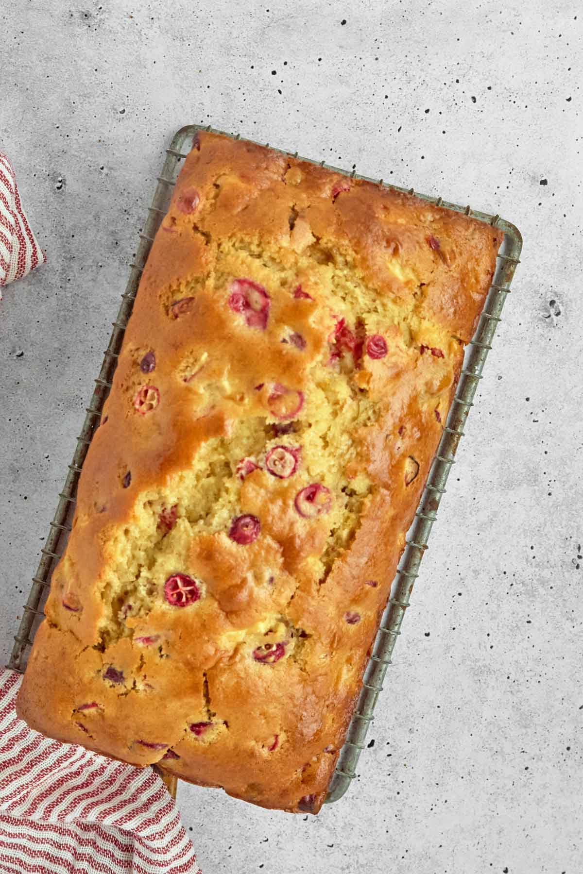 A golden-brown cranberry loaf sits on a wire cooling rack, with visible cranberries throughout the bread and a striped towel nearby-perfect for pairing with easy sheetpan pancakes at your next brunch.