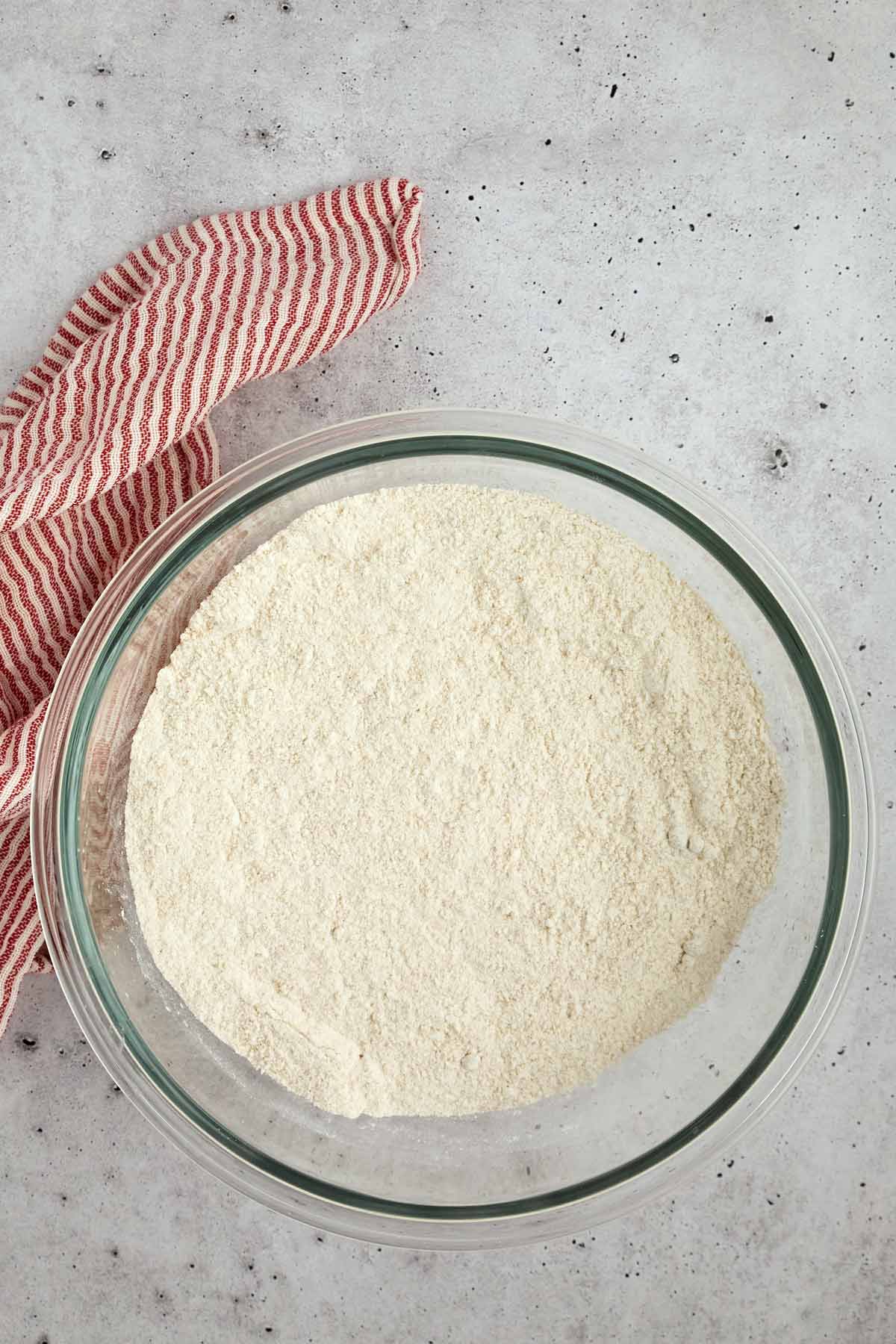 A glass bowl filled with flour sits on a light gray countertop next to a red and white striped cloth-just what you need to start your favorite sheet pan pancakes from mix.