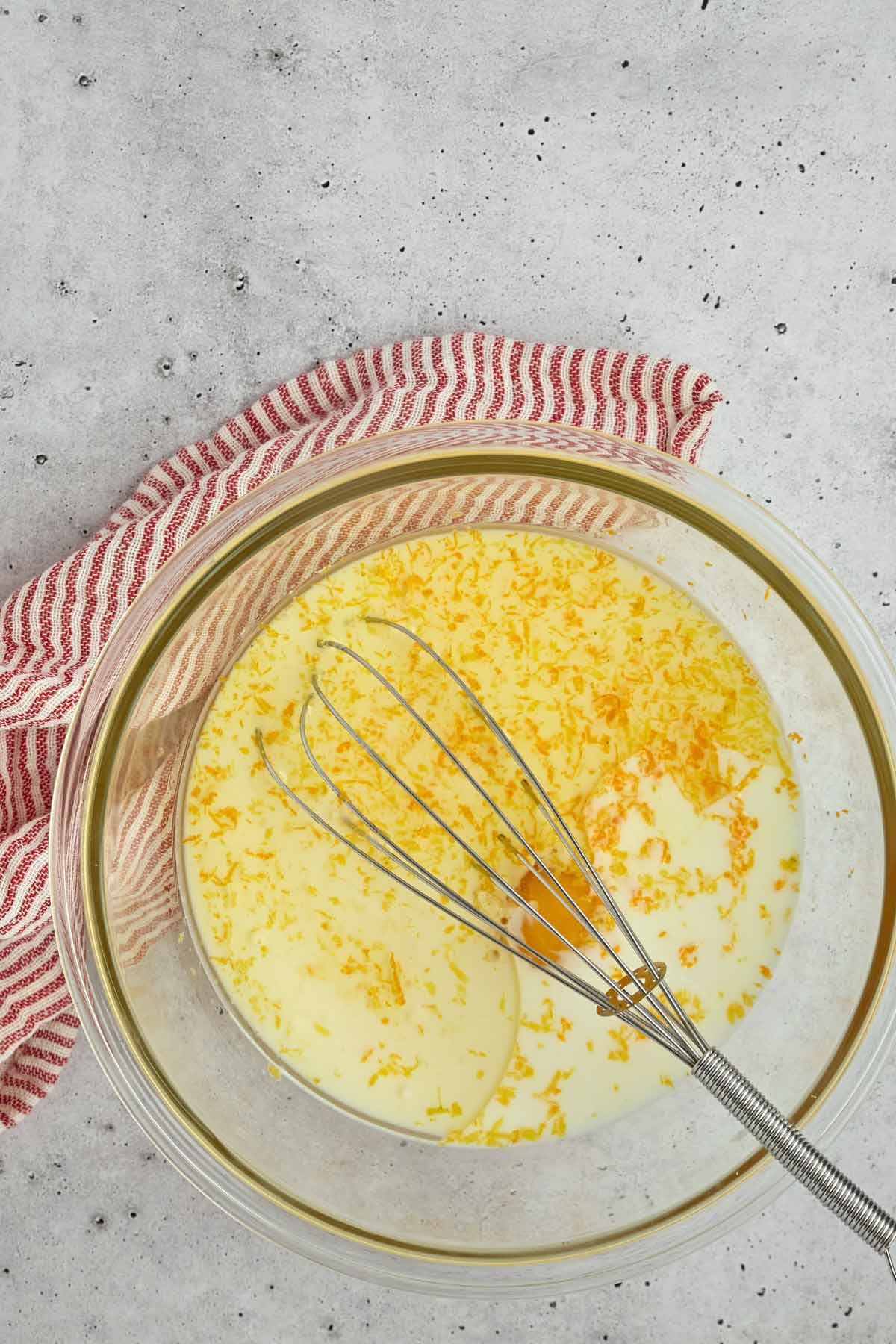 A glass mixing bowl with milk and orange zest, a whisk inside, sits on a gray surface next to a red and white striped cloth-perfect for prepping sheet pan pancakes from mix.