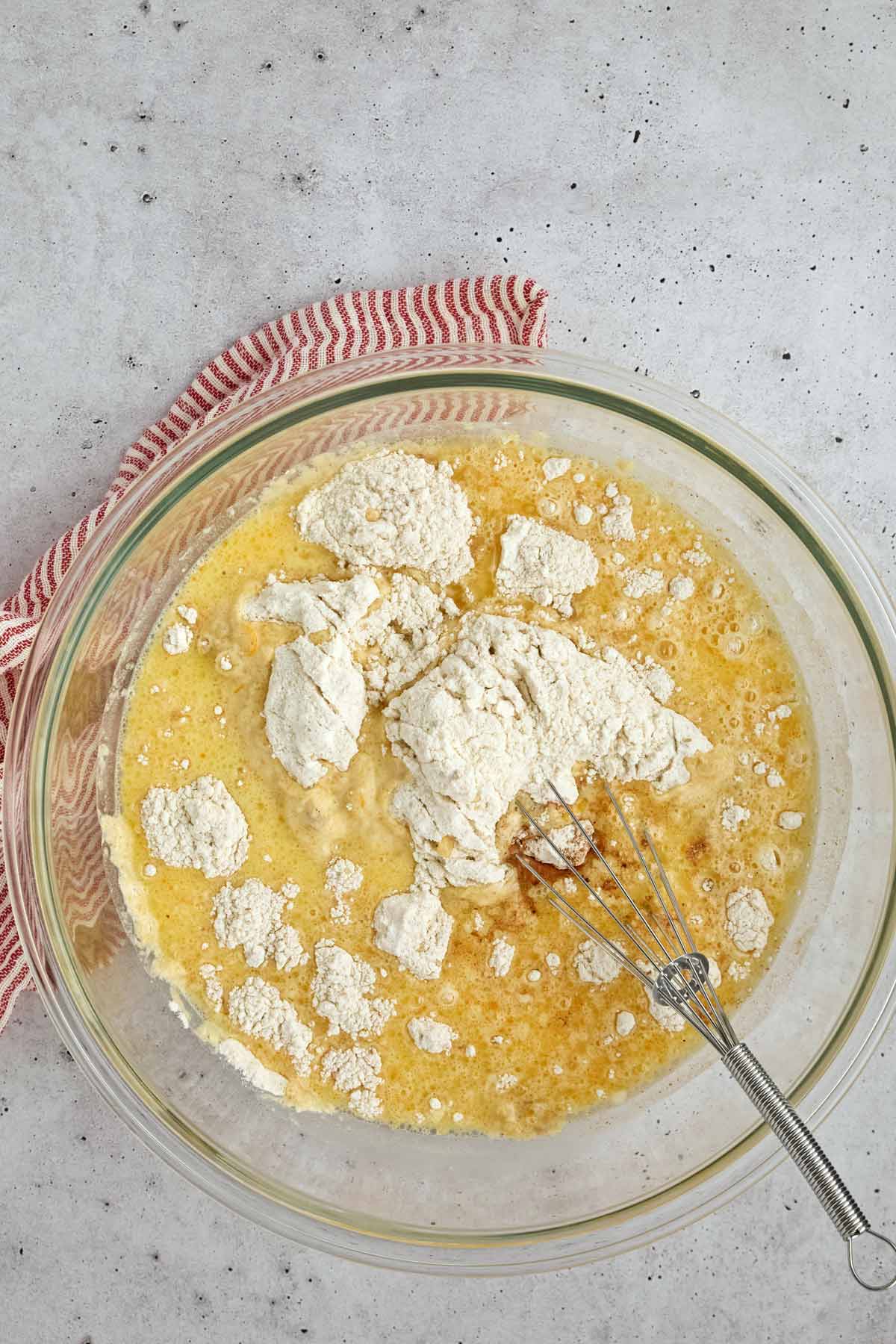 A glass bowl with flour, eggs, and liquid being mixed with a whisk-perfect for making easy sheetpan pancakes-sits on a light surface with a red striped towel beside it.