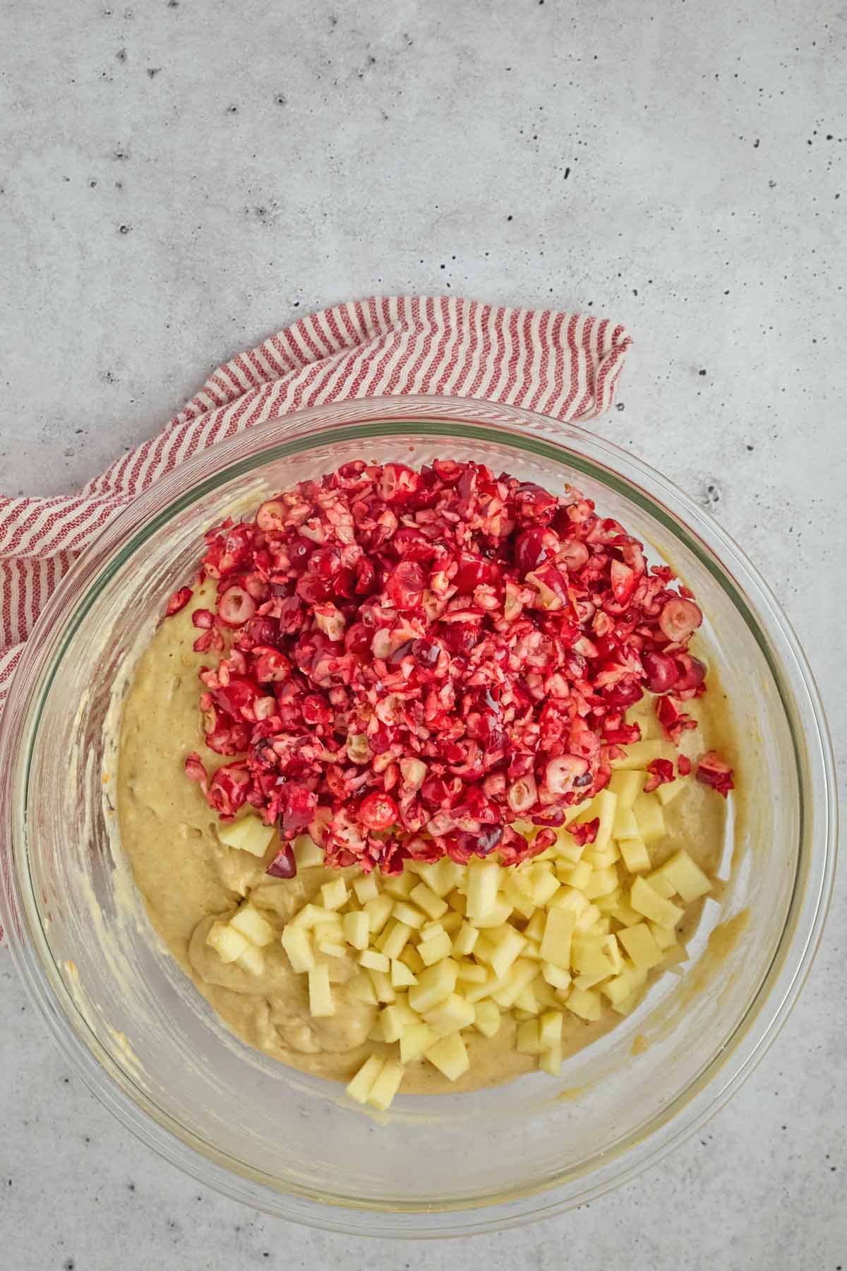 A glass bowl with chopped cranberries and diced apples on top of yellow batter, perfect for making festive sheet pan pancakes, set on a light countertop with a red and white striped cloth nearby.