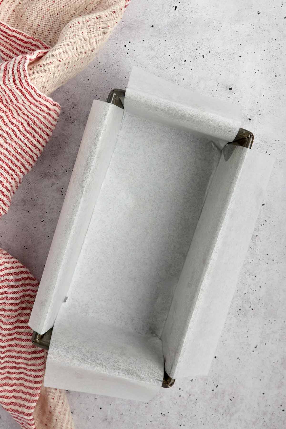 A rectangular baking pan lined with parchment paper sits on a light gray surface next to a red and white striped kitchen towel, ready for your favorite sheetpan pancake recipe.