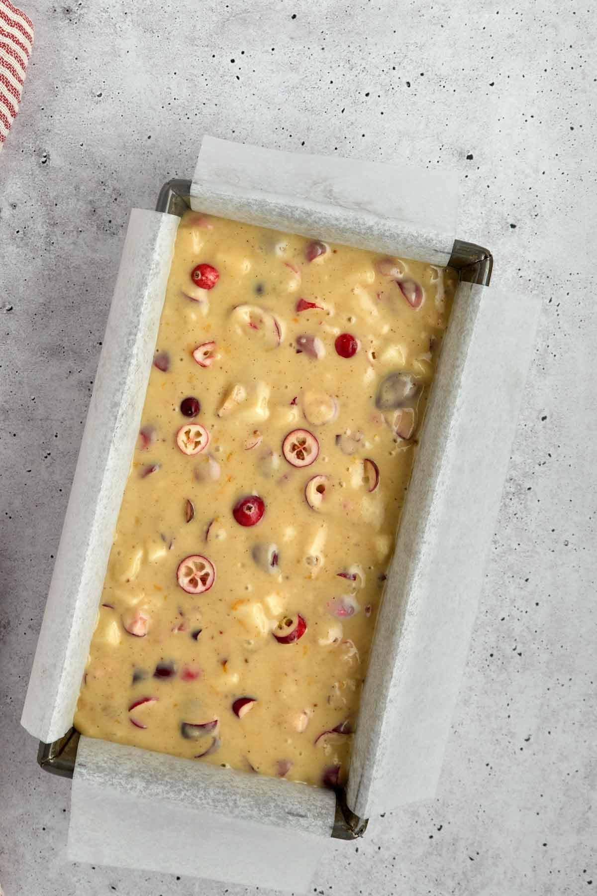 A loaf pan lined with parchment paper filled with unbaked batter containing visible cranberries and nuts sits on a gray surface, inspired by the flavors of a classic sheetpan pancake recipe.