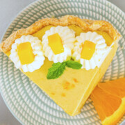 A slice of Mango Pie with a flaky crust, topped with whipped cream, mango cubes, and a mint leaf, is served on a patterned plate with an orange slice—a perfect showcase for your favorite mango pie recipe.