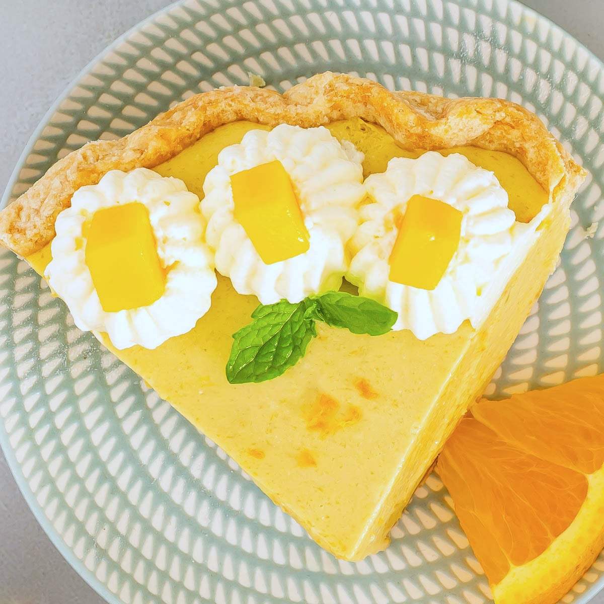 A slice of Mango Pie with a flaky crust, topped with whipped cream, mango cubes, and a mint leaf, is served on a patterned plate with an orange slice-a perfect showcase for your favorite mango pie recipe.