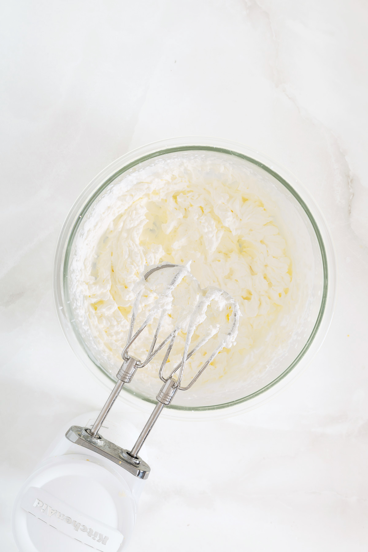 A glass bowl of whipped cream with an electric hand mixer resting on the edge, viewed from above on a white surface-perfect for topping your favorite mango pie recipe.