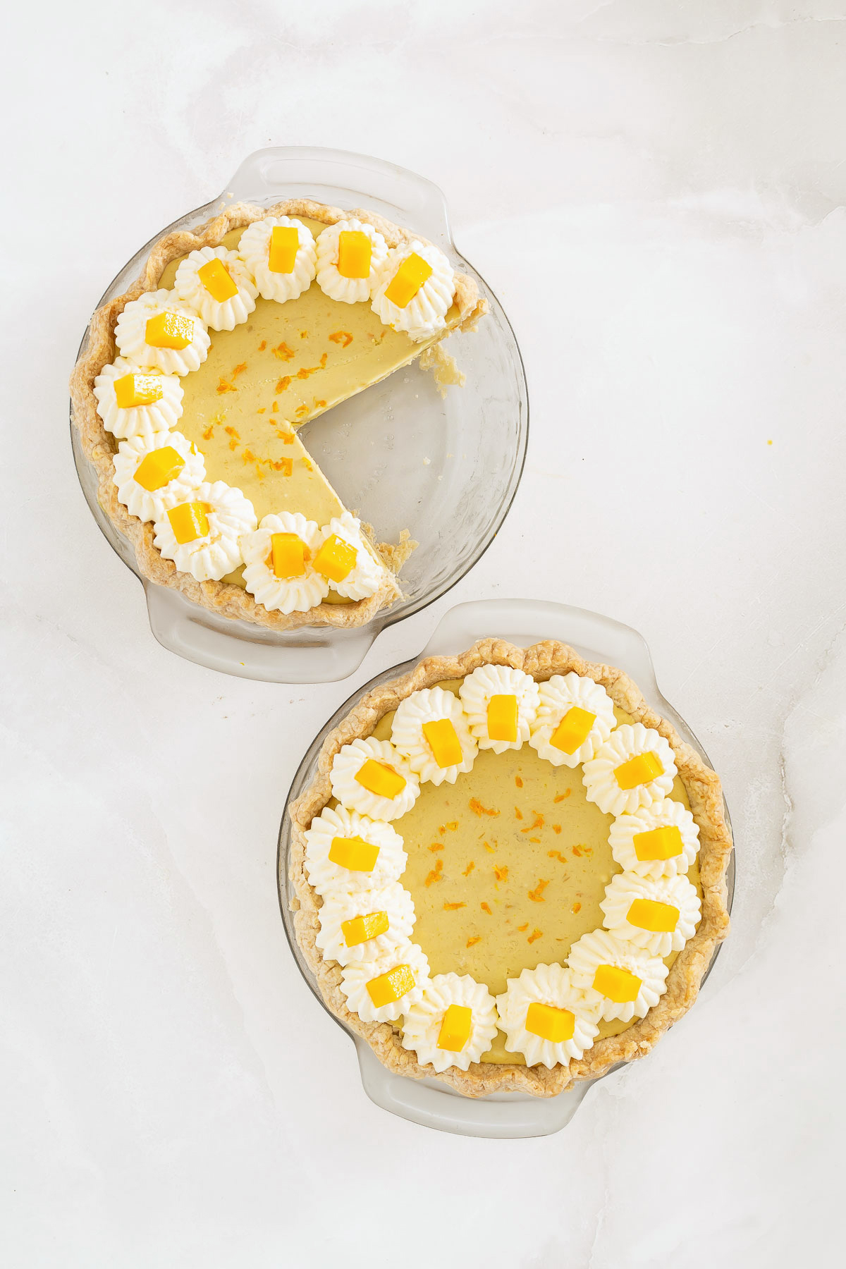 Two lemon pies in glass dishes on a white surface; one pie has a slice missing. Both are topped with whipped cream, small yellow lemon decorations, and sit beside a delicious mango fruit pie for the perfect dessert trio.