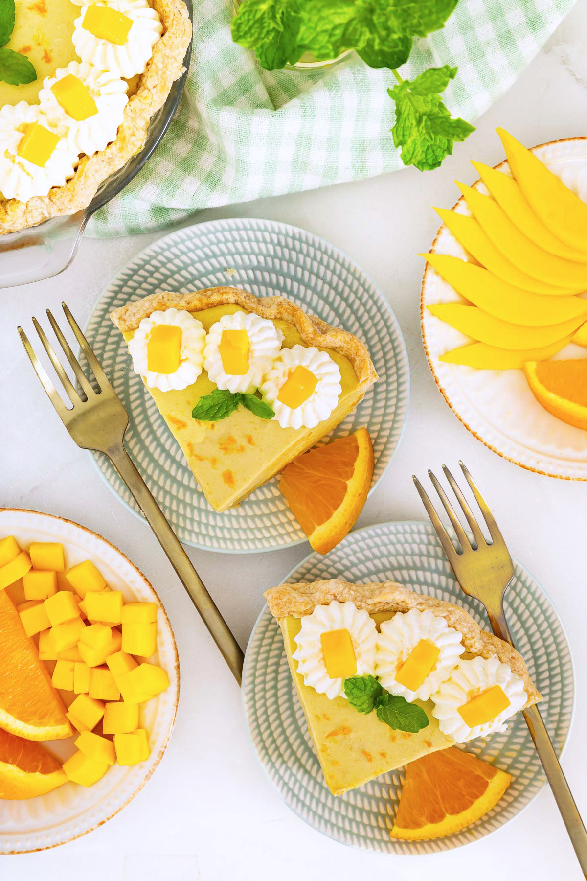 Two plates with slices of mango pie topped with whipped cream, fresh mango pieces, and mint, each served with an orange slice. Surrounding plates hold sliced mango and orange—perfect for trying your favorite mango pie recipe.