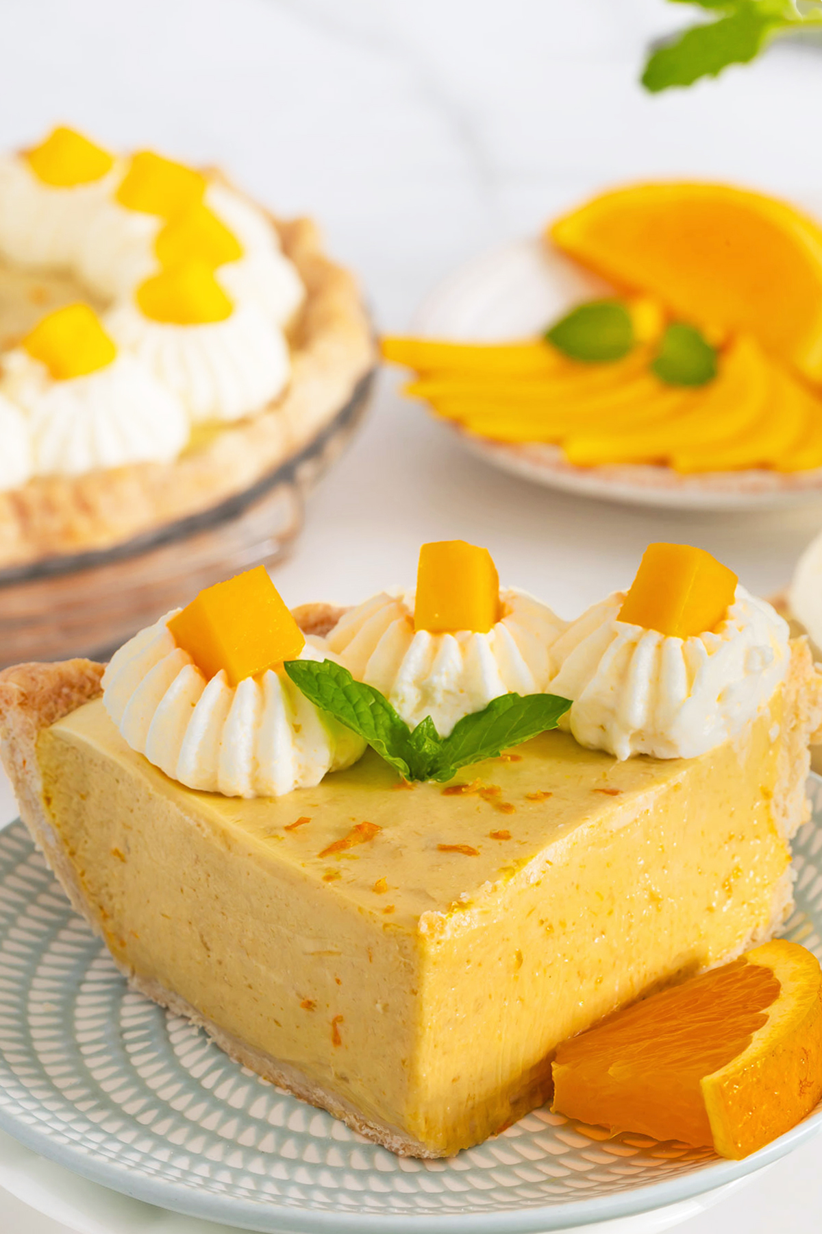 A slice of Mango Pie topped with whipped cream and fresh mango pieces is served on a plate, with a full mango fruit pie and sliced mango in the background.
