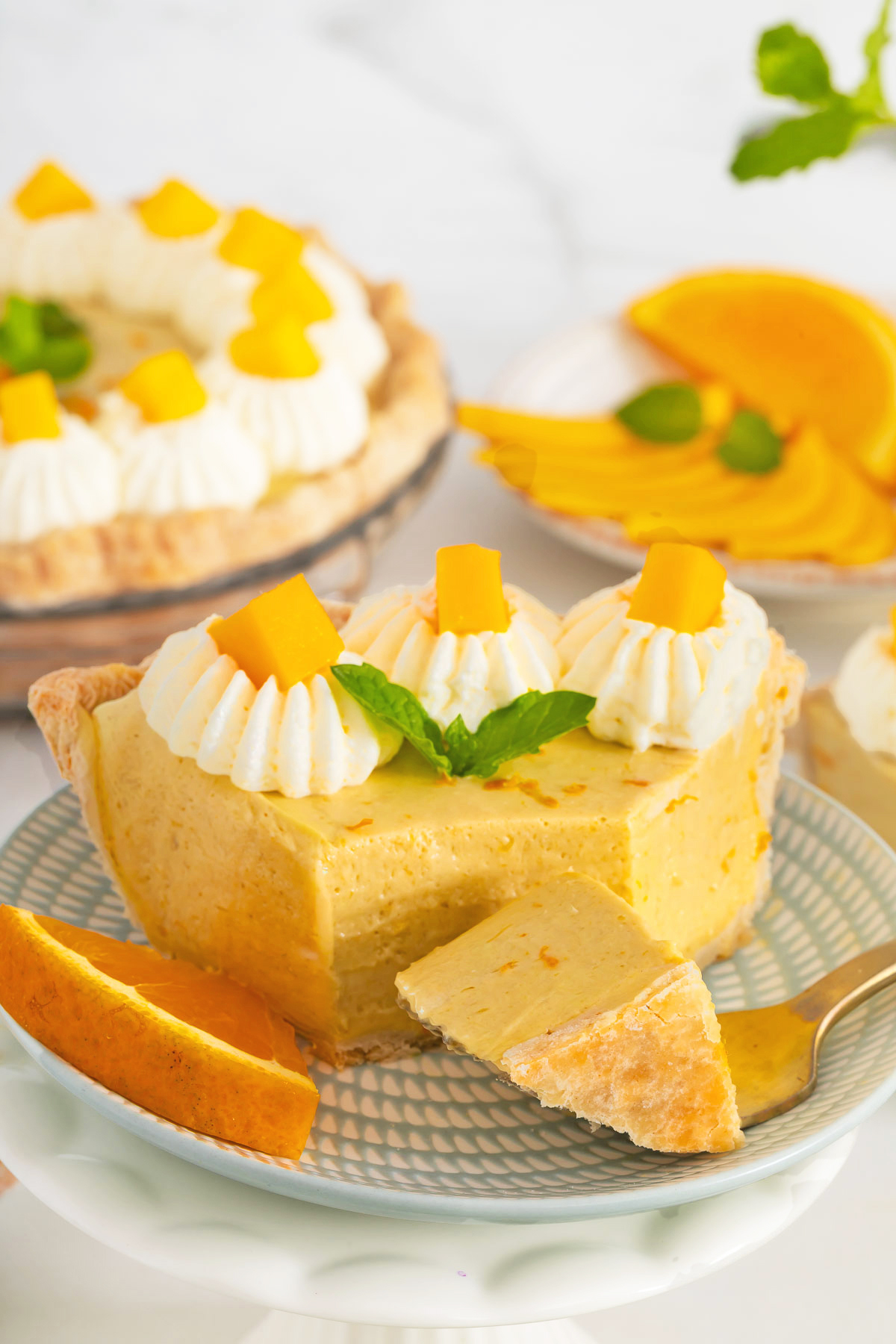 A slice of mango fruit pie topped with whipped cream, mango chunks, and mint leaves sits on a plate with a fork; more pie and sliced mango are visible in the background. Try this delicious mango pie recipe today!.