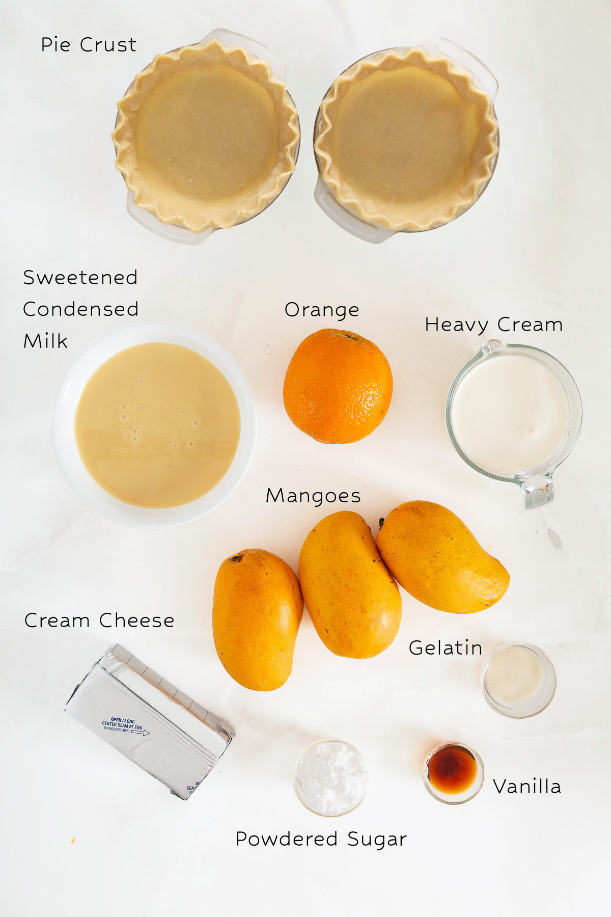 Top-down view of labeled ingredients for a Mango Pie, including pie crust, sweetened condensed milk, orange, heavy cream, mangoes, cream cheese, gelatin, powdered sugar, and vanilla. Perfect for your next mango pie recipe!.