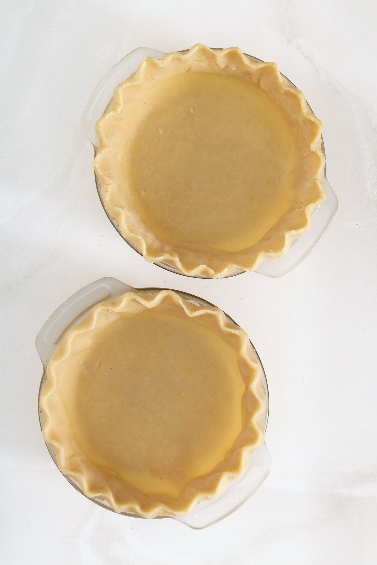 Two unbaked pie crusts with crimped edges in clear glass pie dishes on a white surface, perfect for creating your favorite mango pie recipe.