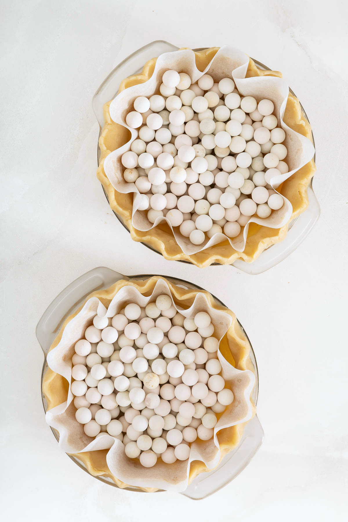 Two unbaked pie crusts lined with parchment paper and filled with ceramic pie weights, ready for your favorite mango pie recipe, are viewed from above on a white surface.