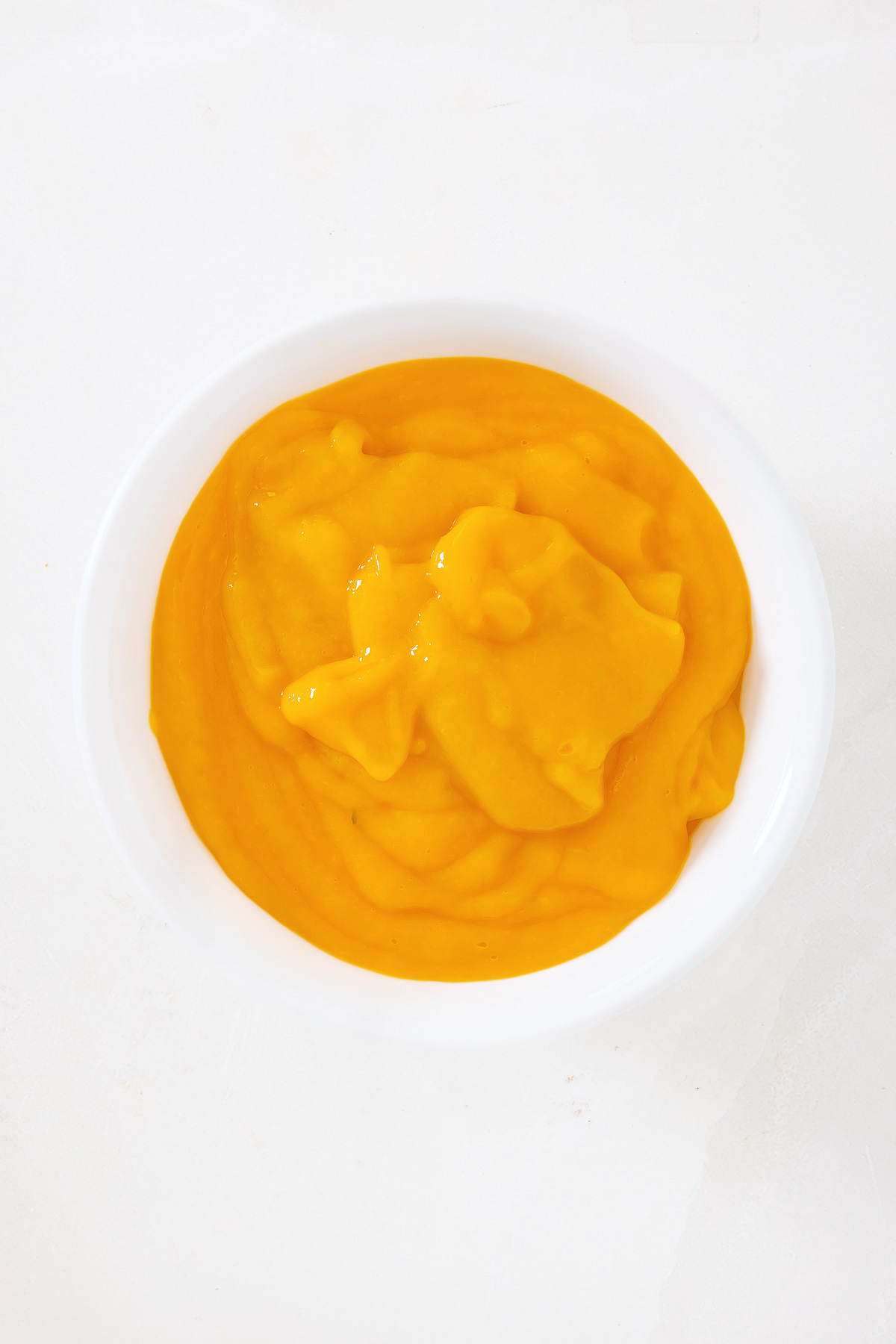 A white bowl filled with smooth, bright orange puree, reminiscent of the vibrant flavors found in a classic mango pie recipe, placed on a white surface.