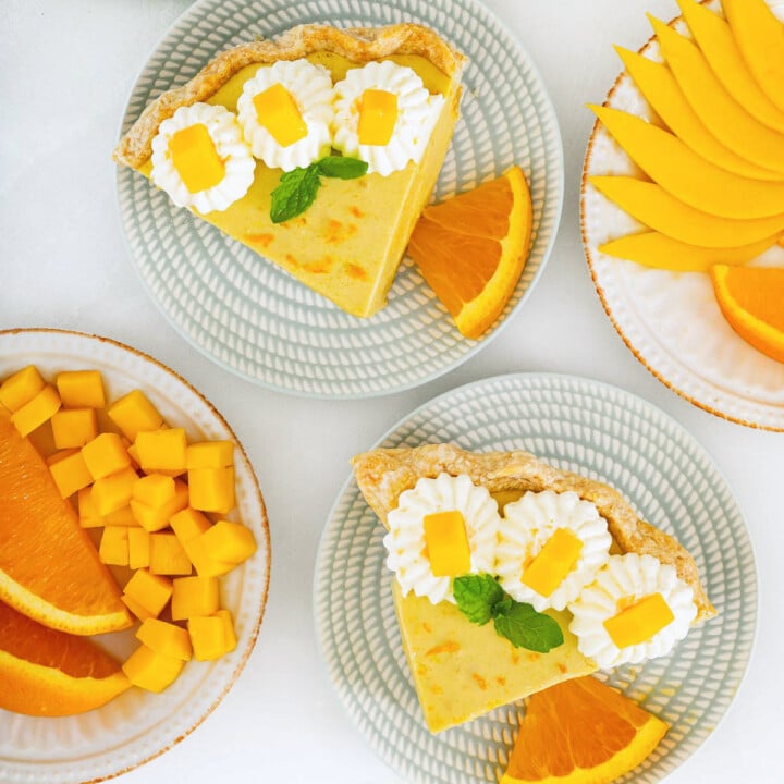 Two slices of pie topped with whipped cream and mango cubes, served with orange slices on plates; additional plates hold sliced mango and orange.