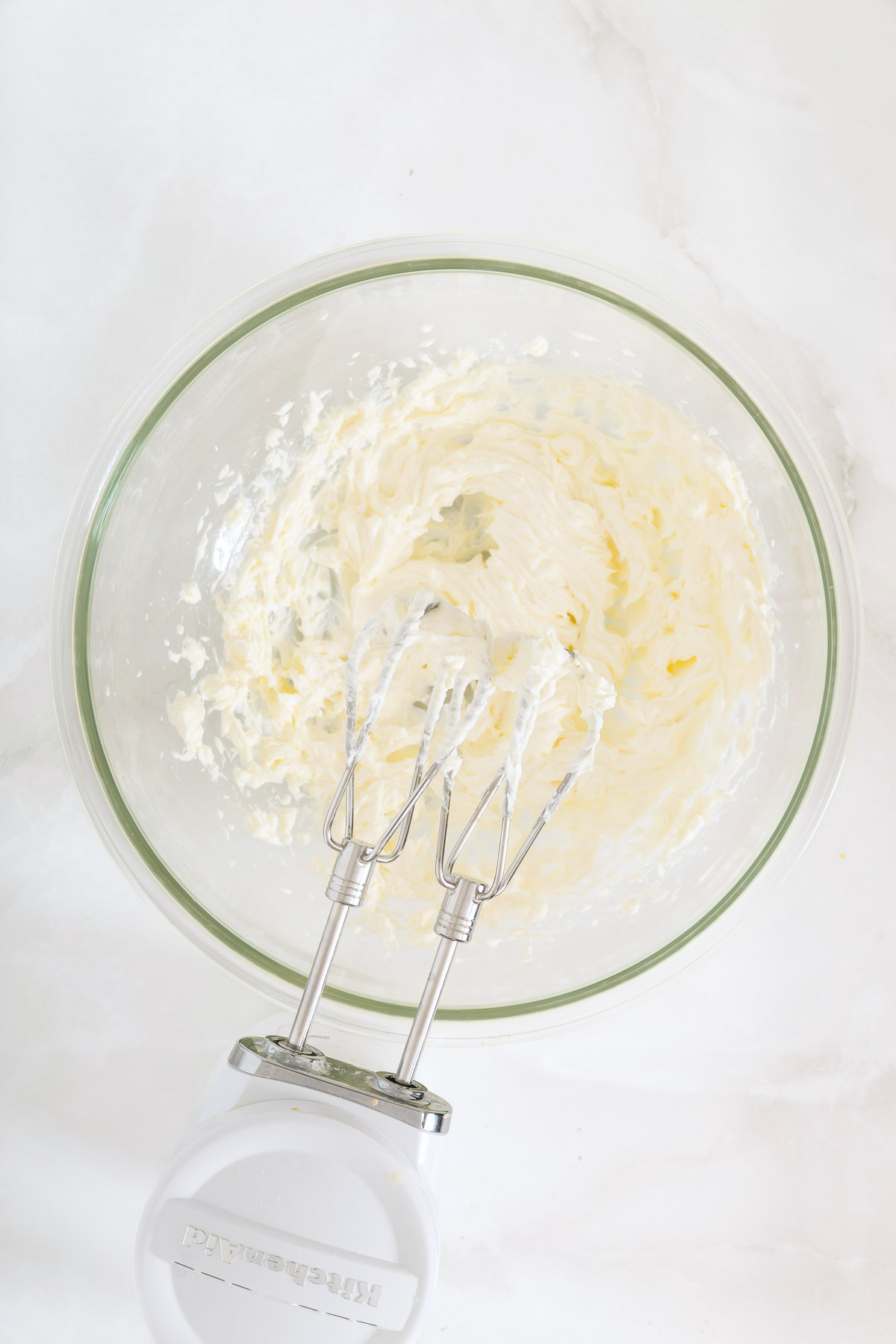 A glass bowl with creamy mixture for Mango Pie being whipped by an electric hand mixer on a white surface, perfect for your next recipe mango pie adventure.