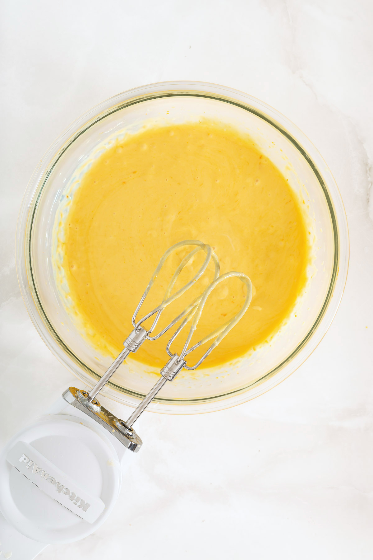 A glass bowl filled with yellow cake batter for a delicious mango pie and a hand mixer with two beaters resting inside on a white surface.