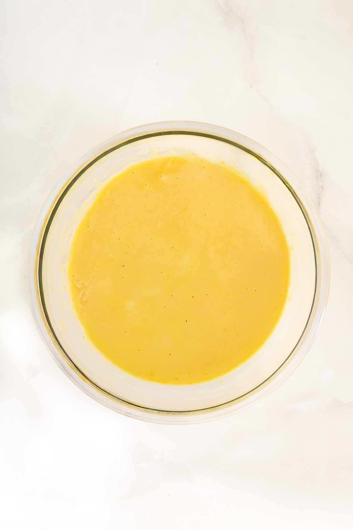 A glass bowl filled with smooth yellow batter for a delicious mango fruit pie sits on a white marble surface.