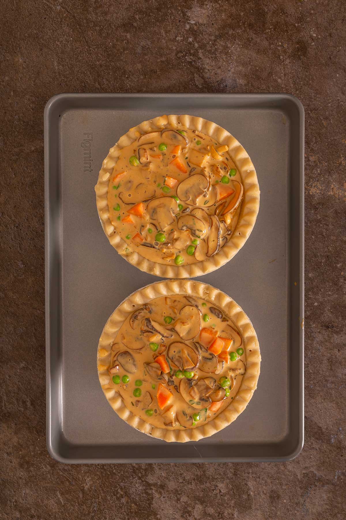 Two unbaked pot pies filled with vegetables and creamy sauce sit on a metal baking sheet atop a brown surface, similar to how you'd prepare an easy sheetpan pancake before baking.