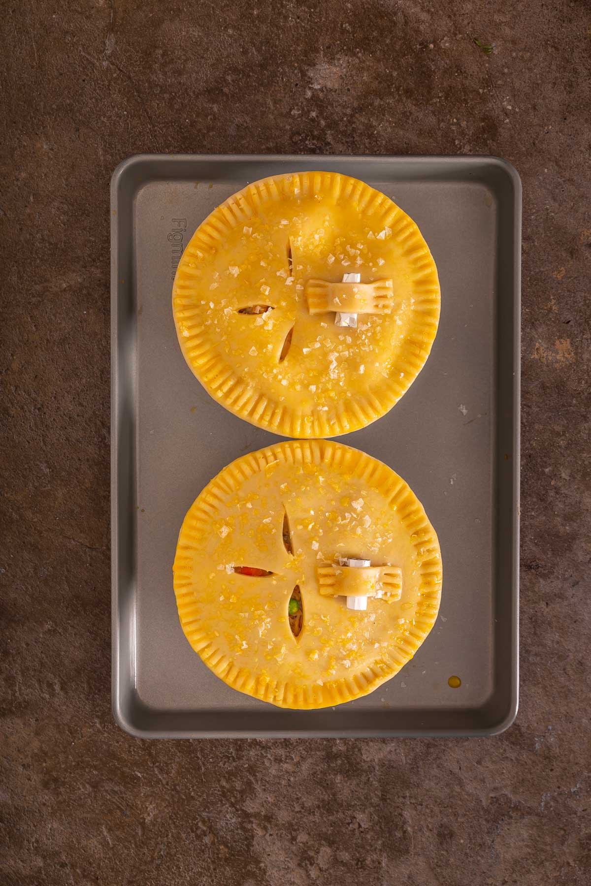 Two unbaked, round savory pies with crimped edges and slits on top sit on a baking sheet, sprinkled with coarse salt-perfect to bake alongside your favorite sheet pan pancakes for a delicious brunch spread.