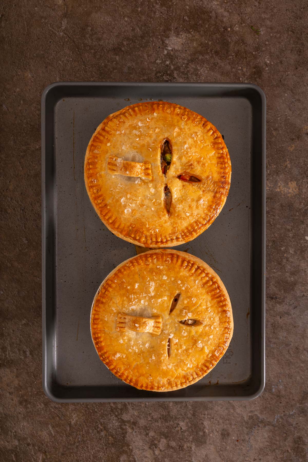 Two golden-brown pot pies with vent slits on top are placed side by side on a rectangular baking tray, reminiscent of the simplicity of an easy sheetpan pancake recipe, set against a stone-like background.