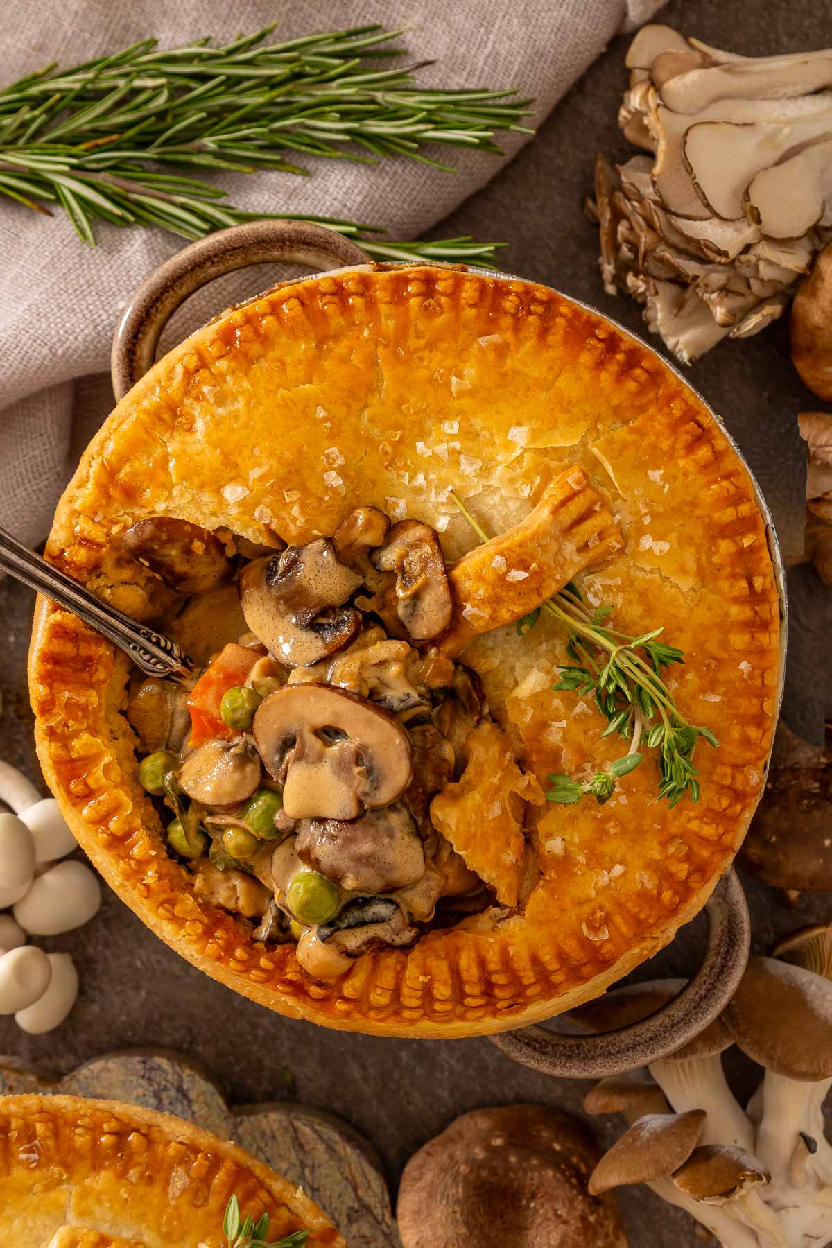 A golden-brown pot pie filled with mushrooms, peas, and carrots sits on a table, partially cut open with a silver spoon inserted, surrounded by fresh herbs and assorted mushrooms-perfect comfort food like easy sheetpan pancakes.