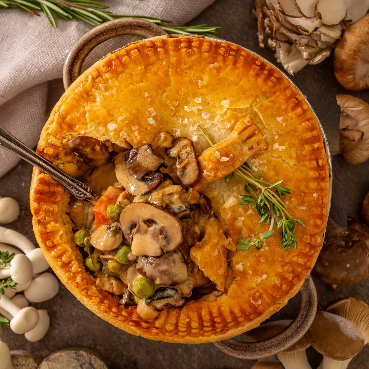 A baked pot pie with a golden crust, filled with mushrooms, peas, and carrots. Like easy sheetpan pancakes, it's simple to prepare and perfect for sharing. A spoon rests inside, and fresh herbs garnish the dish.