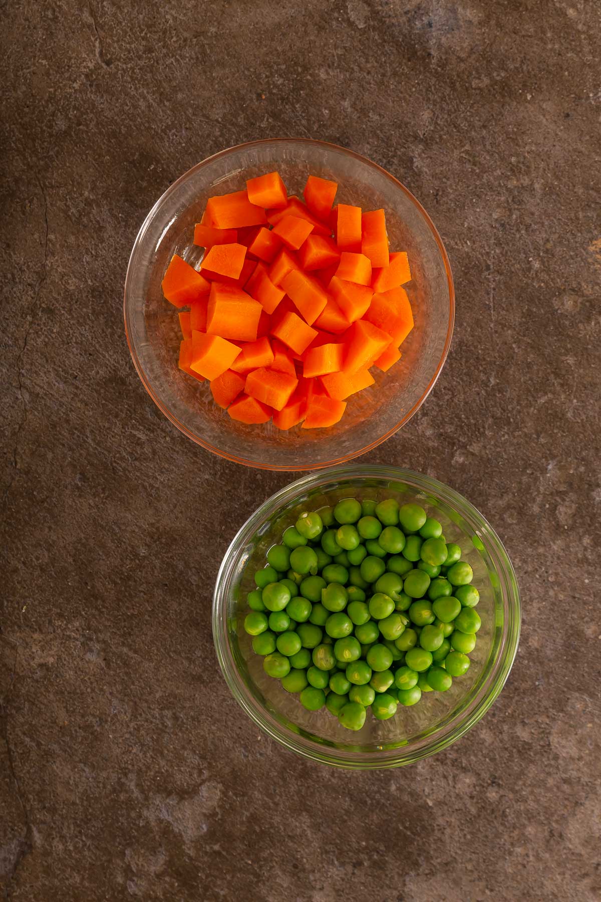 Two glass bowls on a brown surface, one with diced carrots and the other filled with green peas-perfect veggies to serve alongside easy sheetpan pancakes for a balanced meal.