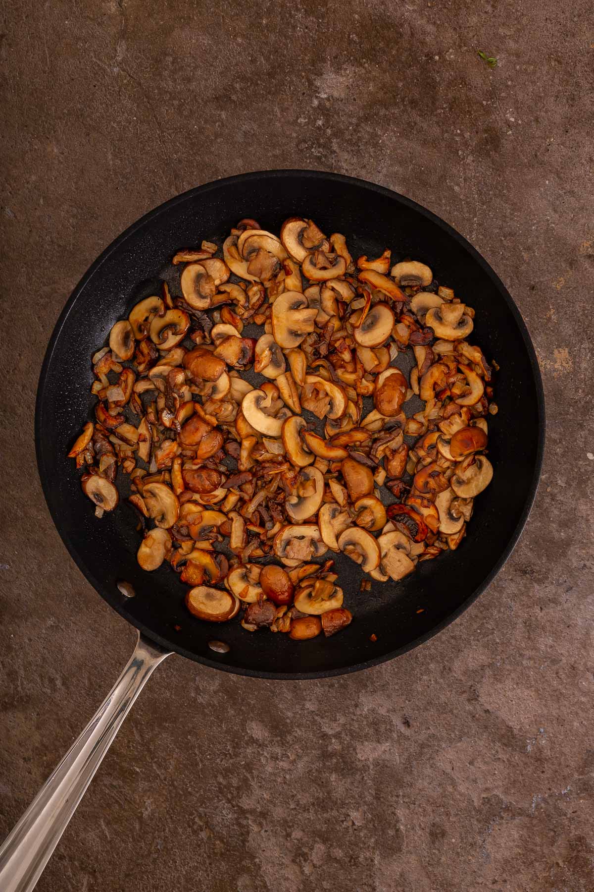 A black skillet filled with cooked, sliced mushrooms sits on a brown, textured surface-perfect as a savory topping for your favorite sheetpan pancake recipe.