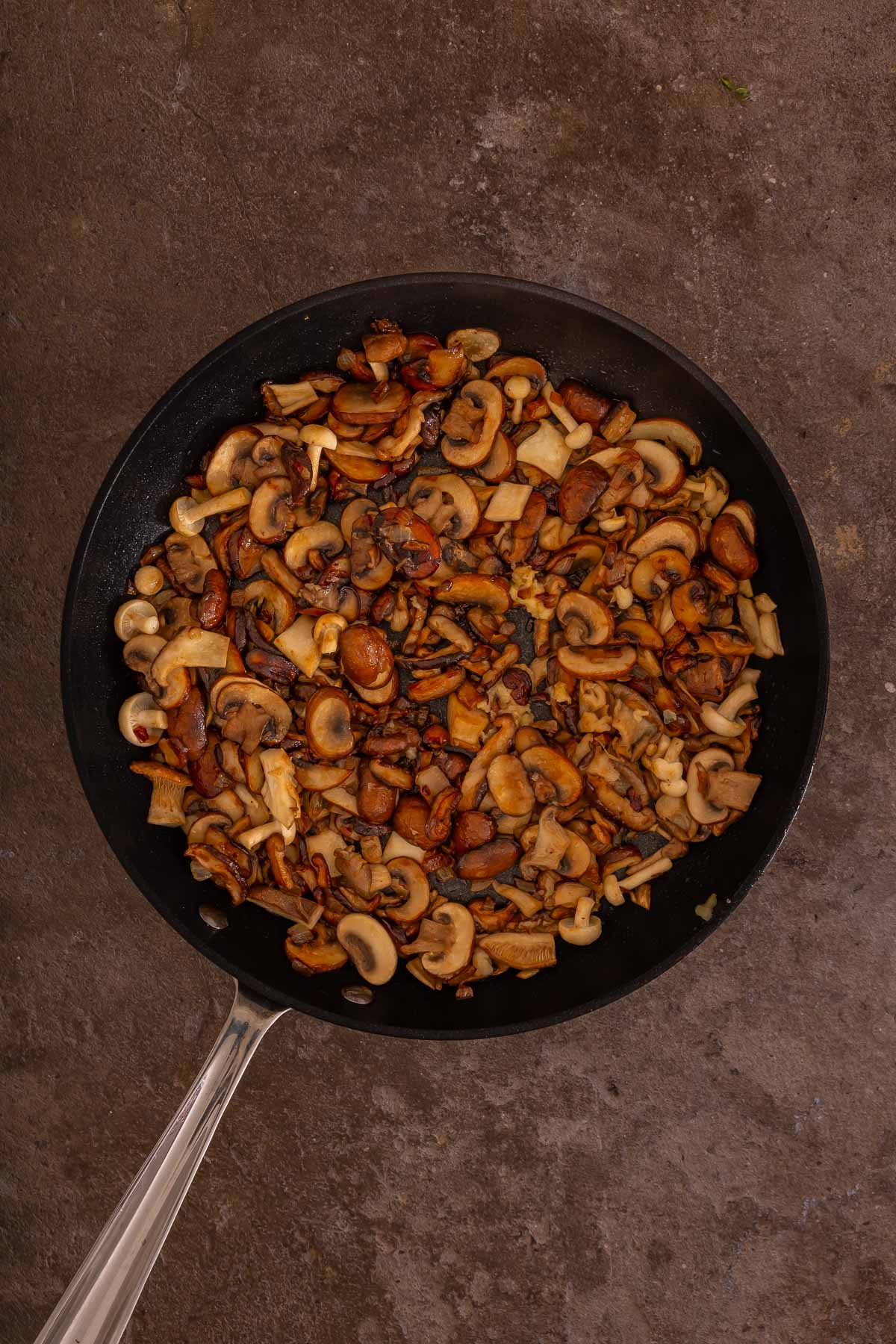 A frying pan filled with sautéed mixed mushrooms sits on a textured brown surface, ready to accompany your favorite breakfast dishes like fluffy sheetpan pancakes.