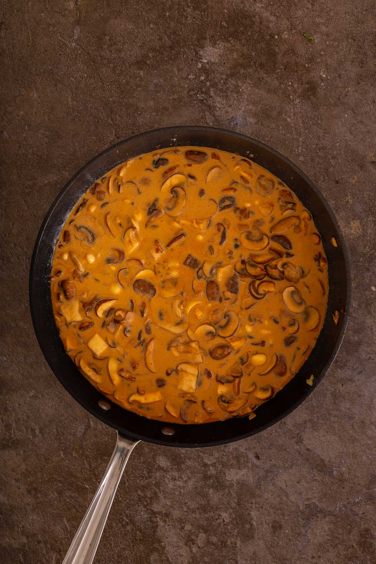 A black skillet filled with creamy mushroom sauce, featuring sliced mushrooms in a light brown, thick liquid, sits on a dark textured surface-perfect for pairing alongside your favorite easy sheetpan pancakes.