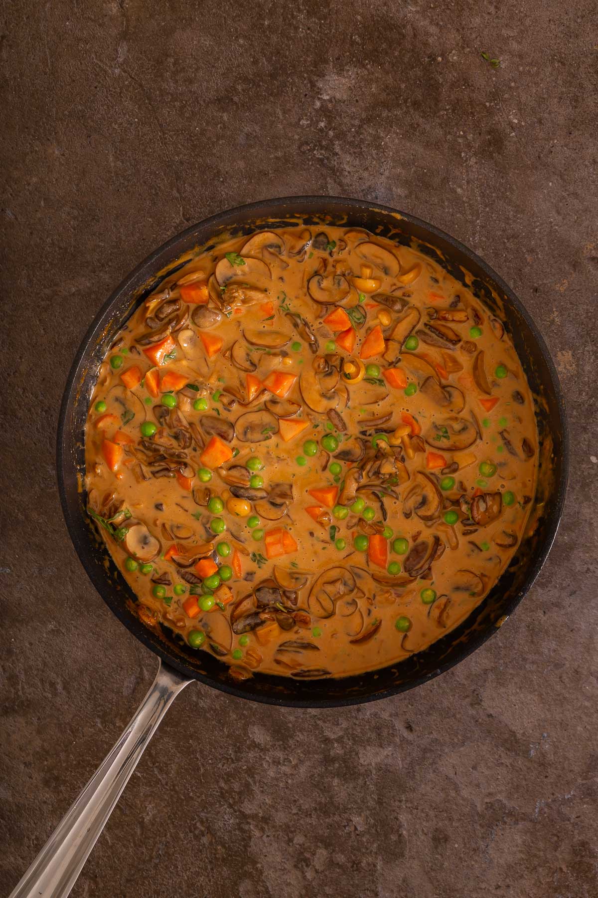 A pan filled with creamy mushroom stew containing sliced mushrooms, diced carrots, and green peas sits on a brown countertop beside a delicious sheetpan pancake recipe, perfect for a hearty meal.