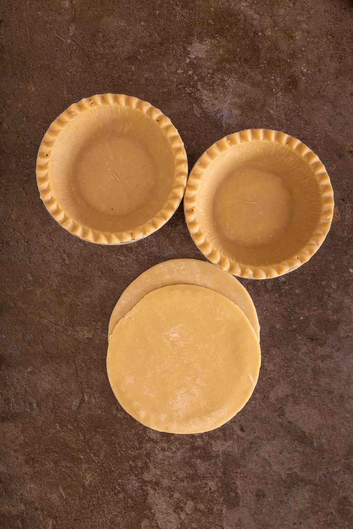 Three unbaked pie crusts in tins and two flat rounds of pie dough are arranged on a brown surface, perfect for baking pies or even trying a creative twist like making a sweet sheetpan pancake recipe.