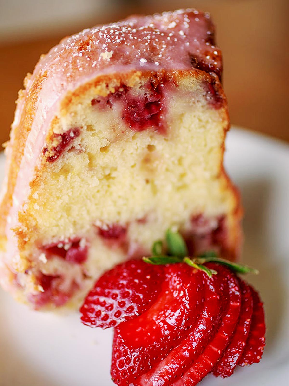 A slice of strawberry cake with pink glaze sits on a plate, reminiscent of the beloved strawberry spoon cake recipe, garnished with a fan of fresh sliced strawberries in front.
