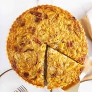 A baked onion tart with one slice being served, showing a golden filling with visible onion and bacon pieces, placed on a white surface with a plate and fork nearby—perfect for anyone who enjoys comforting brunch dishes, similar to the ease of an easy sheetpan pancakes recipe.