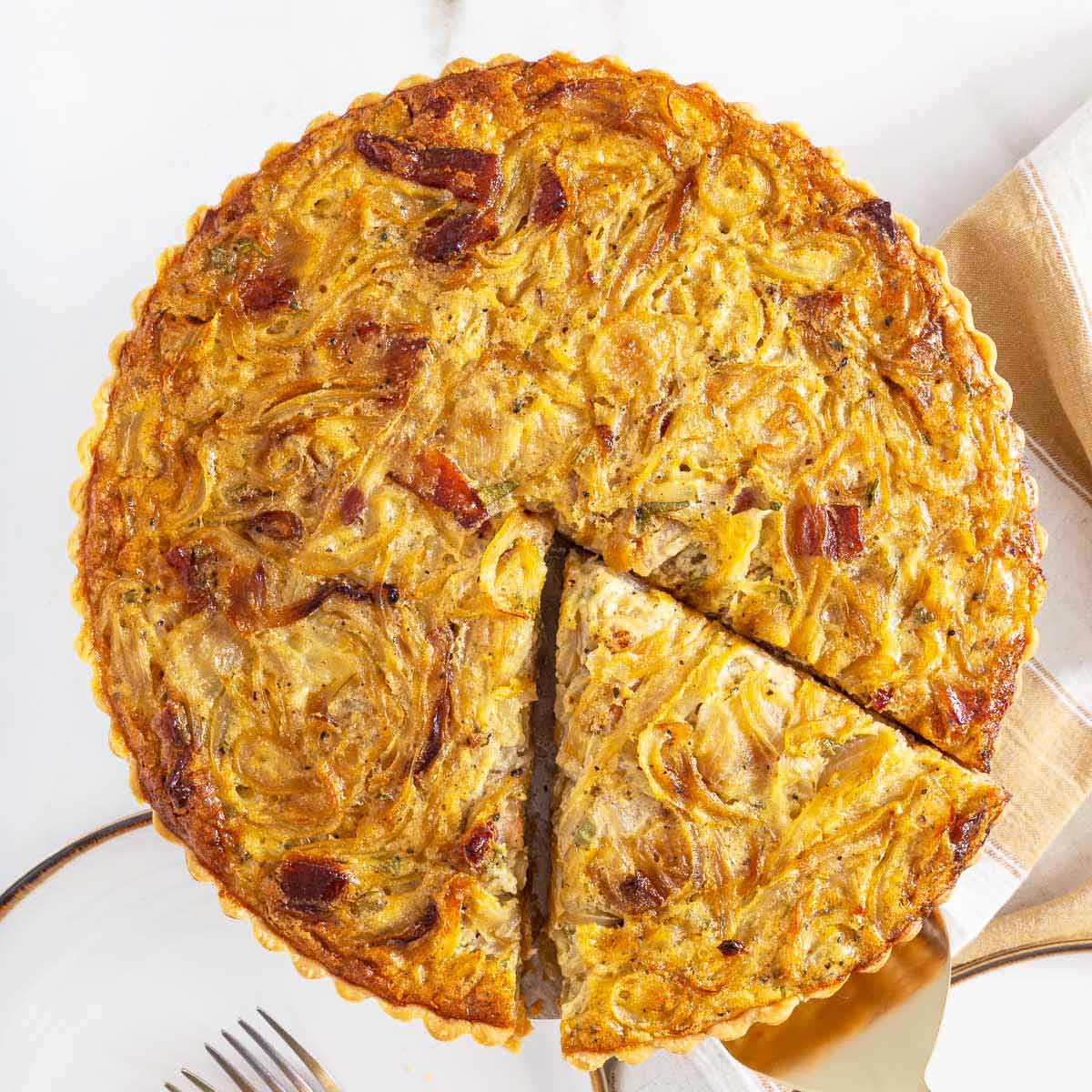 A baked onion tart with one slice being served, showing a golden filling with visible onion and bacon pieces, placed on a white surface with a plate and fork nearby-perfect for anyone who enjoys comforting brunch dishes, similar to the ease of an easy sheetpan pancakes recipe.