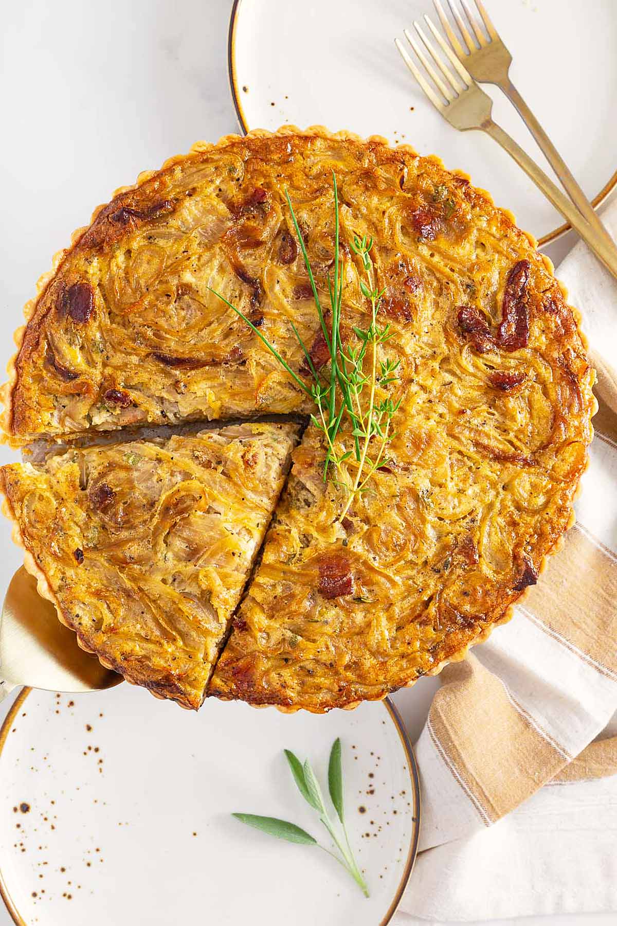 A golden-brown quiche with one slice removed, garnished with fresh herbs, sits on a plate with gold utensils and a beige napkin nearby—reminiscent of the inviting presentation often seen with easy sheetpan pancakes.