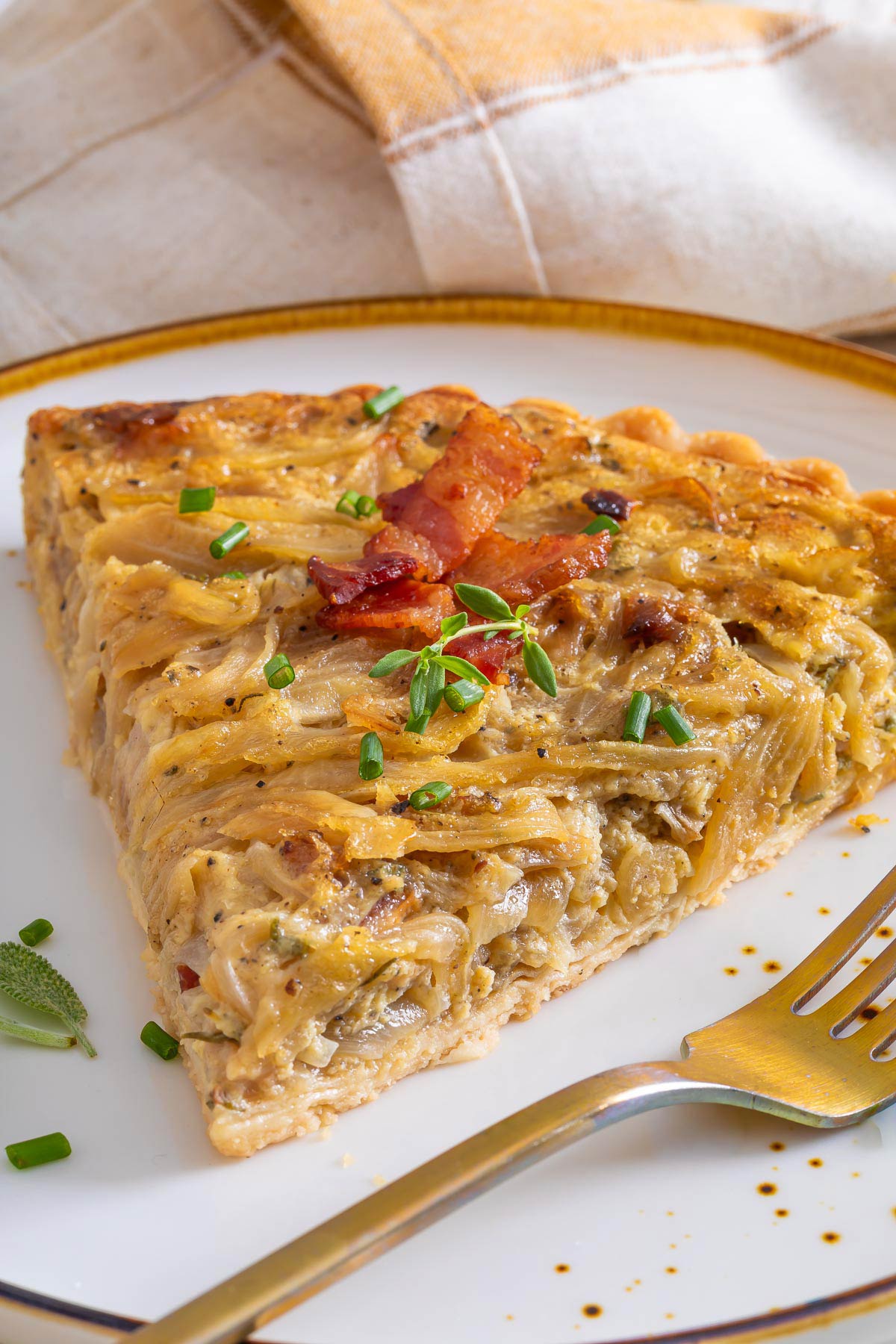 A slice of savory onion tart topped with bacon and chopped chives sits elegantly on a white plate with a gold rim, next to a fork—a delightful alternative to sweet breakfasts like sheetpan pancake recipes.