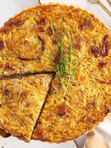 A baked onion tart with a golden crust, garnished with fresh herbs, sits on a plate beside a pie server and partially removed slice-its tempting aroma reminiscent of a savory twist on your favorite sheetpan pancake recipe.
