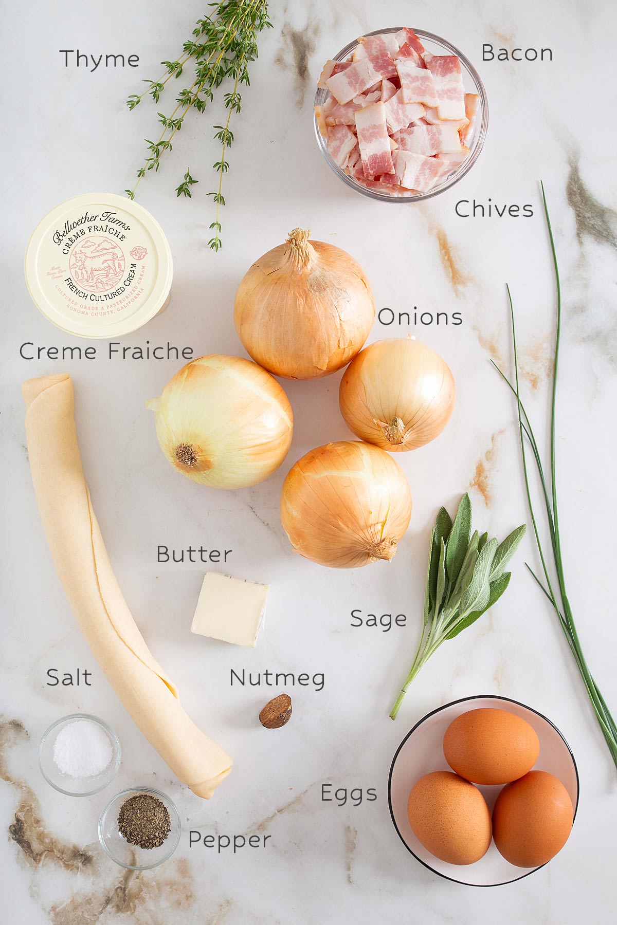 Ingredients for an easy sheet pan pancake recipe: thyme, bacon, chives, creme fraiche, onions, butter, sage, eggs, nutmeg, salt, pepper, and a sheet of pastry-all beautifully arranged on a marble surface.