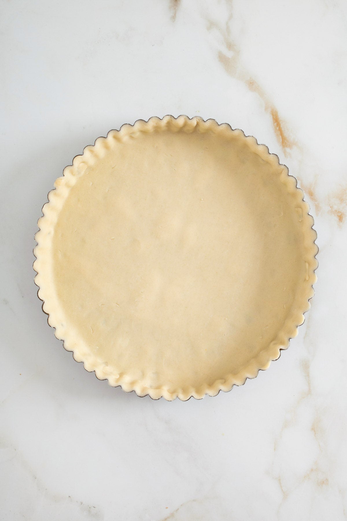 Unbaked pie crust evenly pressed into a round, fluted tart pan on a white marble surface-much like preparing sheetpan pancake recipe batter before spreading it to bake.