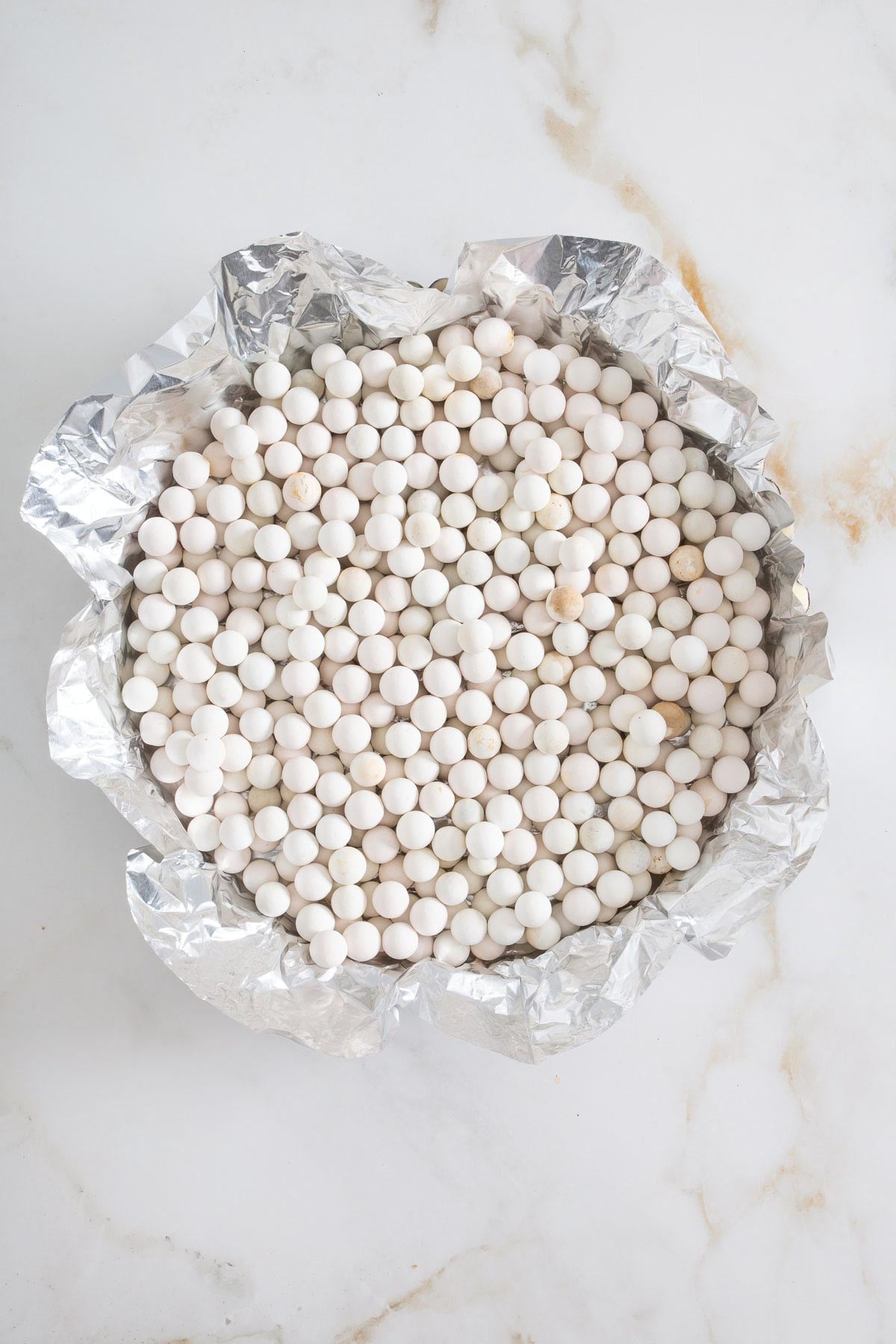 A pie tin lined with crumpled aluminum foil is filled with white ceramic pie weights, set on a white marble surface-just the kind of preparation you'd use before baking a flaky crust for an easy sheetpan pancakes recipe.