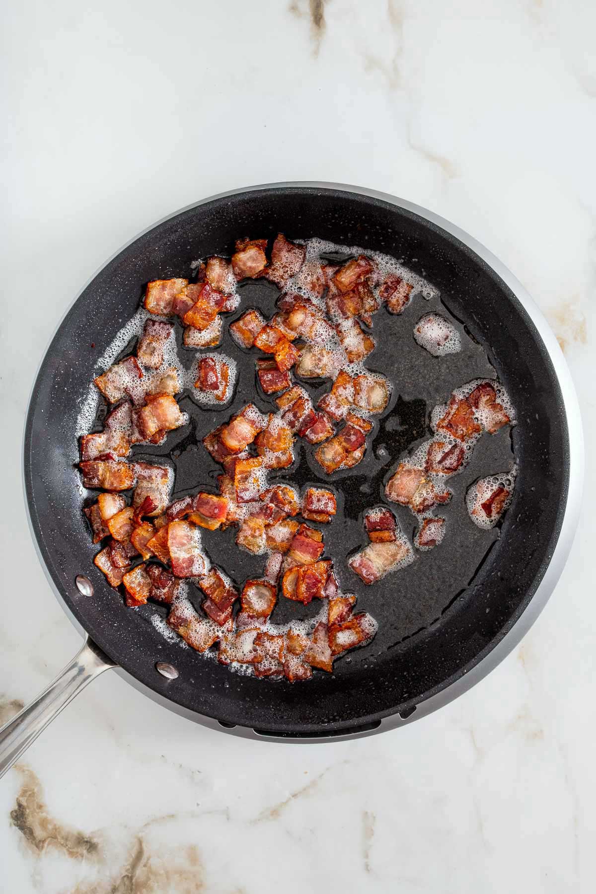 Chopped bacon pieces cooking and sizzling in a black frying pan on a marble countertop, perfect for topping your favorite sheetpan pancake recipe.