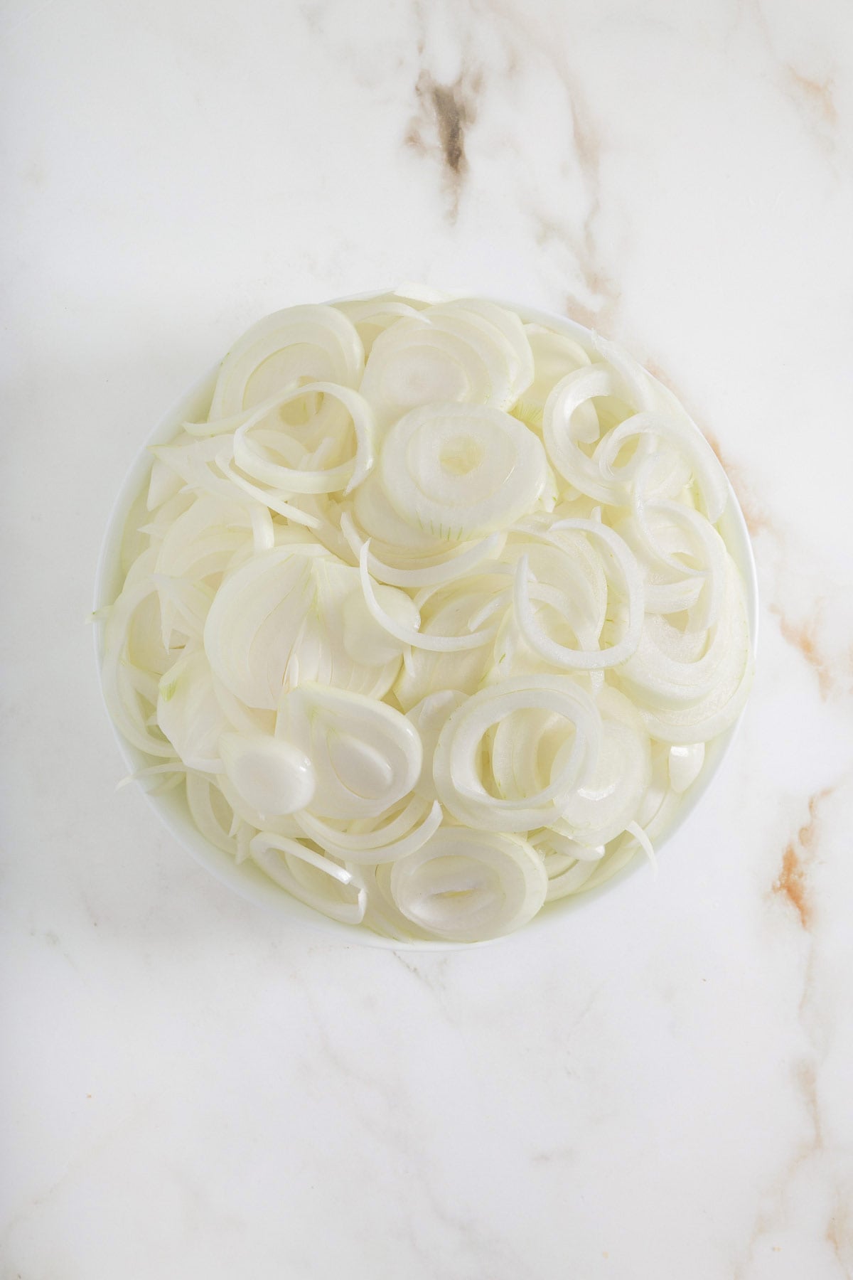 A white plate filled with sliced raw white onions, placed on a white marble surface-perfect for topping a savory sheetpan pancake or complementing your favorite sheet pan pancakes from mix.