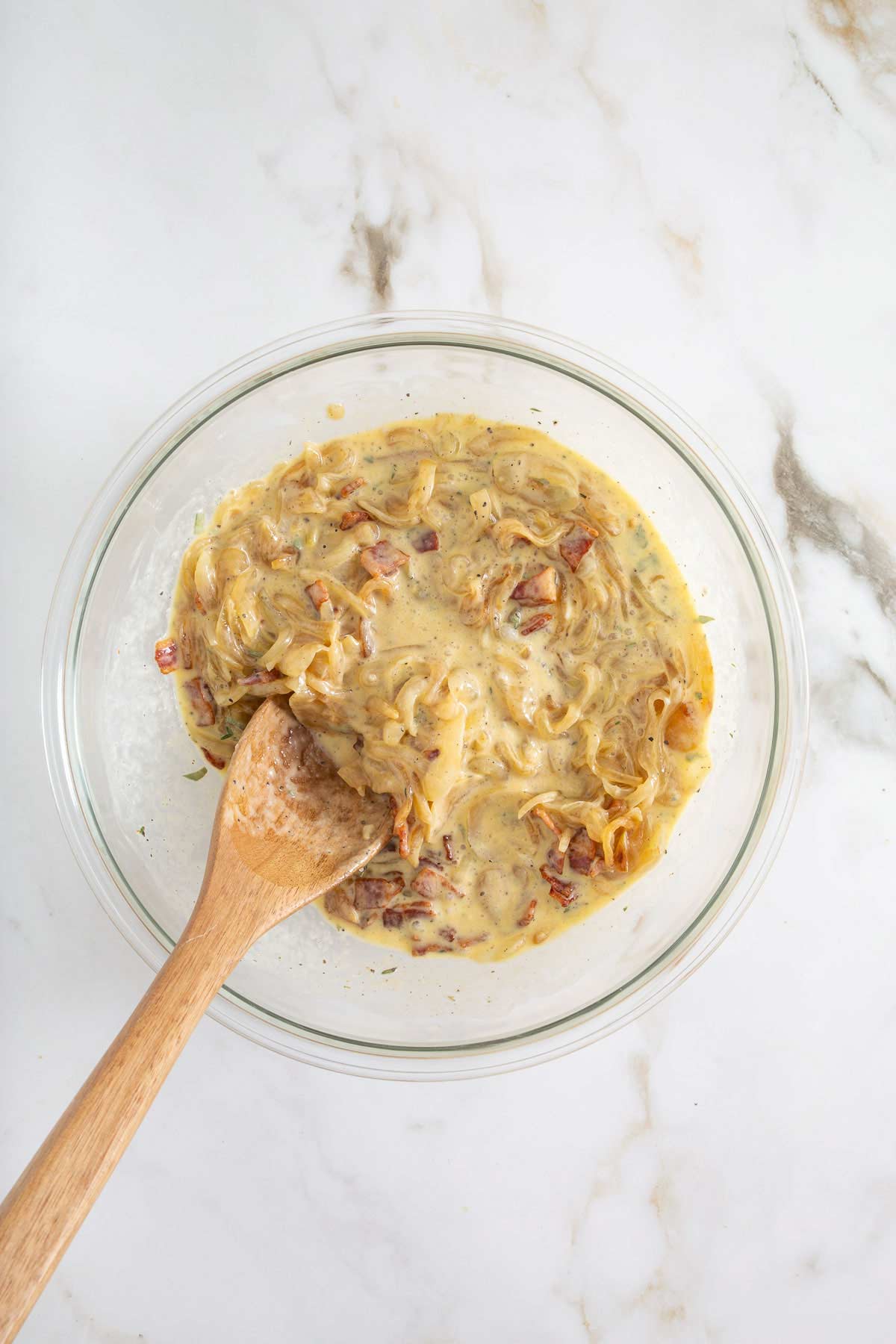 A glass bowl with a creamy onion mixture and chopped bacon, stirred with a wooden spoon, sits on a white marble surface-perfect for adding a savory twist to your easy sheetpan pancakes.