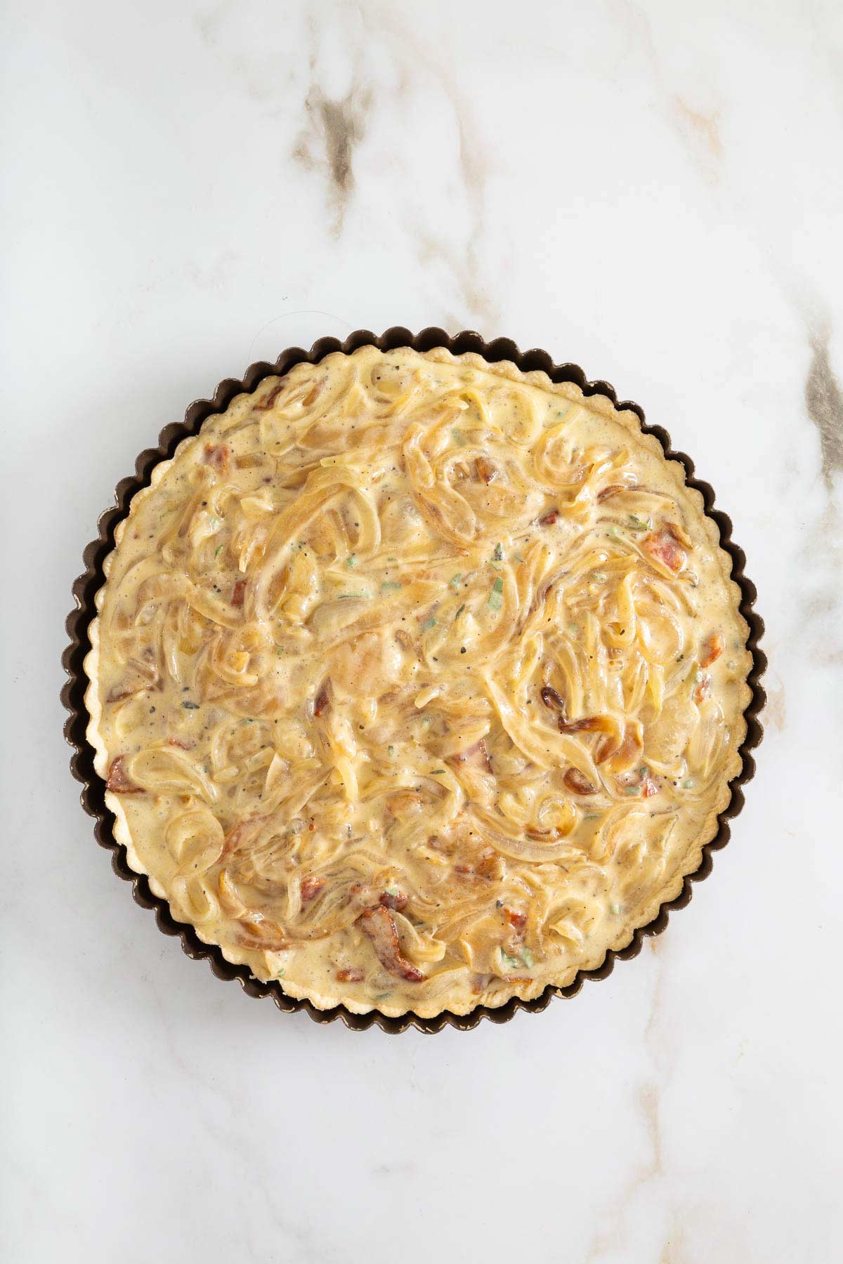 Unbaked savory tart filled with a creamy mixture of onions and herbs in a round tart pan, set on a white marble surface-perfect for serving alongside easy sheetpan pancakes at your next brunch.